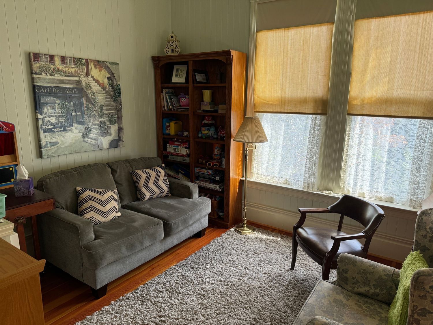 A second room with a gray couch, a bookshelf of games, and tall sunny windows